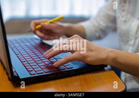 Close-up of woman's hands typing on a laptop keyboard, multitasking with a pen and notepad. Stock Photo