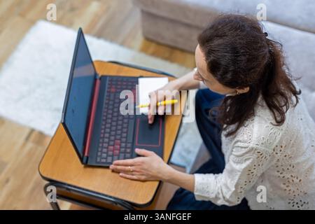 Woman working on laptop at home, taking notes with a pen while focusing on the screen. Stock Photo