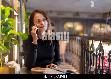 Woman having a phone call at a cafe table, young woman with glasses talking on the phone in a cozy cafe. Stock Photo