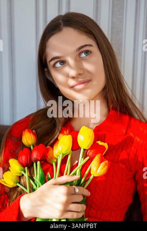 Thoughtful woman holding vibrant tulip bouquet. A young woman in a red sweater gazes thoughtfully while holding a bouquet of red and yellow tulips Stock Photo