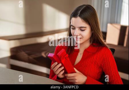 Woman opening a red gift box indoors. A young woman in a red sweater holds and opens a red gift box, expressing curiosity and joy in a cozy room Stock Photo
