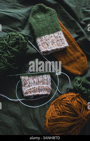 Dark green and brown yarn ball with knitted socks and knitting needles. Wool threads with deep green background. Woven crochet closeup. Knitting hobby Stock Photo