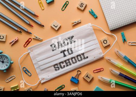 Back to school is written on a white mask. The mask is placed on a table with various office supplies such as pens, pencils, and paper clips Stock Photo
