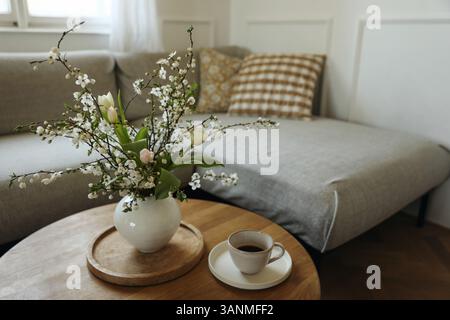 Moody spring interior still life. Cup of coffee. Round wooden table. Tulips blooming cherry plum tree branches bouquet in vase. Cozy linen sofa with Stock Photo
