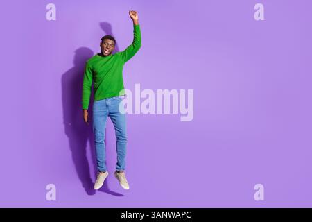 Young man in green sweatshirt jumping joyfully on purple background expressing happiness and excitement. Stock Photo