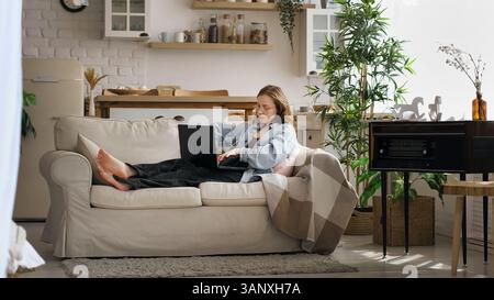 A woman in a denim jacket sits on a couch, focused and thoughtful, holding a notebook and pen while looking at her laptop. This image captures the Stock Photo