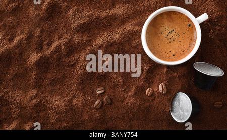 White porcelain espresso cup on a heap of ground coffee with beans and black aluminum capsules around it. Top view. Stock Photo