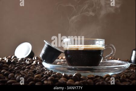 Glass cup of espresso on a clear plate on top of a heap of coffee beans with capsules around it. Front view. Stock Photo