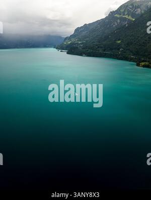 Aerial view of a boat sailing along the Brienzersee Lake coastline in summertime during a rainy day with low clouds, Bonigen, Bern, Switzerland. Stock Photo