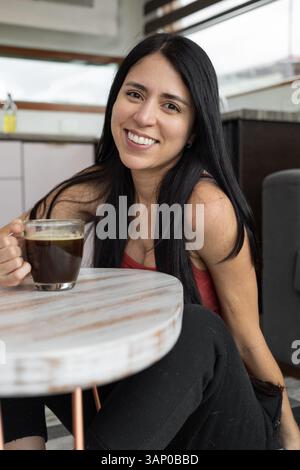 A smiling young woman sips coffee in a cozy café, radiating pure joy and warmth as she enjoys a cheerful moment in a lively setting. Stock Photo