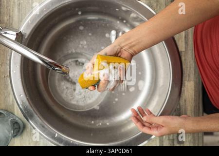Close-up of a woman’s hand squeezing a sponge full of soap while washing dishes in a bright, modern kitchen environment. Stock Photo