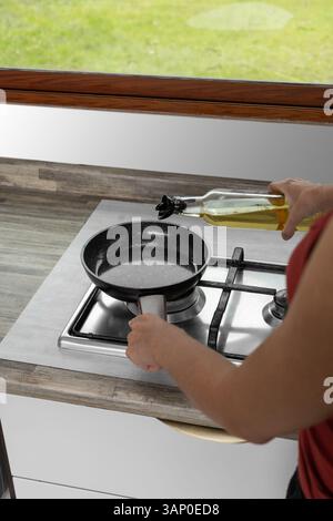 Seen from behind, a woman pours oil into a skillet on a modern stove, with natural light from a window overlooking a garden. Stock Photo