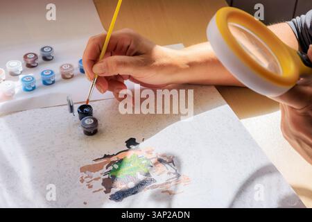 Painting by numbers. Brushes and paints on canvas close-up, woman painting looking at canvas through magnifying glass. High quality photo Stock Photo