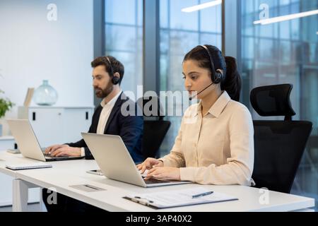 Two call center employees, a woman and a man, are working on laptops at their desks while wearing headsets. Stock Photo