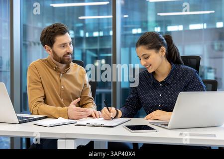 A man and a woman are collaborating in an office setting, with the woman signing documents. They appear to be reviewing a report. Stock Photo