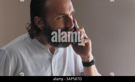 A focused man with a beard is using a smartphone, engaging in a conversation. Wearing a casual white shirt indoors, the scene captures a moment of con Stock Photo