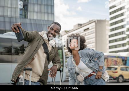 African American tourist couple having fun taking selfies on a busy European city street with modern skyscraper architecture. Stock Photo