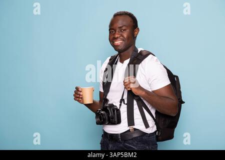 Smiling male model with a backpack and disposable cup looking directly at the camera, exuding confidence and readiness for a new creative project. Black photographer standing with his gear and coffee. Stock Photo