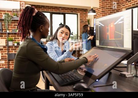 Black female employee sits at desk, updating her asian creative manager on latest progress of the project. Professional women editors collaborate on startup design, sharing ideas in brick wall office. Stock Photo