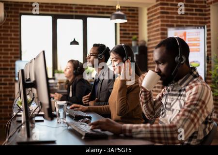 Diverse telemarketers seated at their workstations, communicating with remote clients and handling business requests. Coffee, headsets, and computers emphasizing customer support office environment. Stock Photo