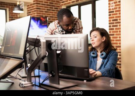 African american man standing and discussing with caucasian woman at desk, analyzing product designs on computer. Diverse employees cooperate in preparing creative projects in brick wall office. Stock Photo
