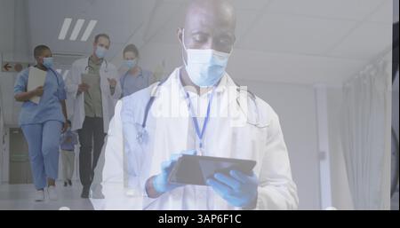 Composite of male doctor in face mask using tablet, and diverse doctors in masks walking in corridor Stock Photo