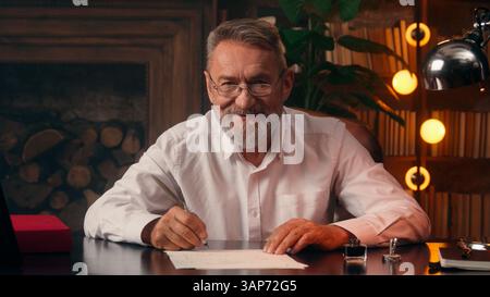 Old Caucasian man writer holding fountain pen paper text writing author journalist work looking at camera smiling male mature pensioner elderly Stock Photo