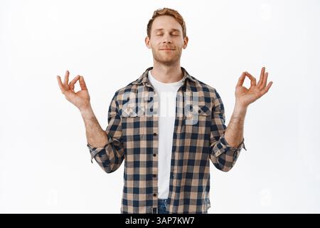 Redhead man close eyes and meditates with zen mudras, smiling and breathing relaxed, peaceful meditation or yoga, trying to calm down, standing over w Stock Photo