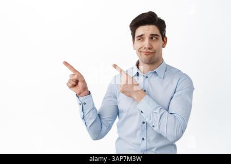 Doubtful businessman smiling skeptical, pointing and looking aside at logo with suspicious hesitant face, standing against white background. Stock Photo