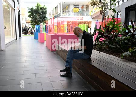 Bearded man looking down at his mobile phone while sitting on a wooden bench in a quiet shopping centre Stock Photo