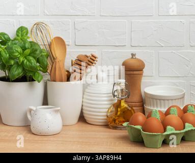 Kitchen counter displaying fresh eggs, basil plant, oil, spices, and various utensils against a white brick wall, embodying a home cooking atmosphere Stock Photo