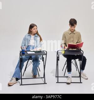 Teen boy and girl sitting at desks in classroom, reading textbooks, focused on study process against grey background. Stock Photo