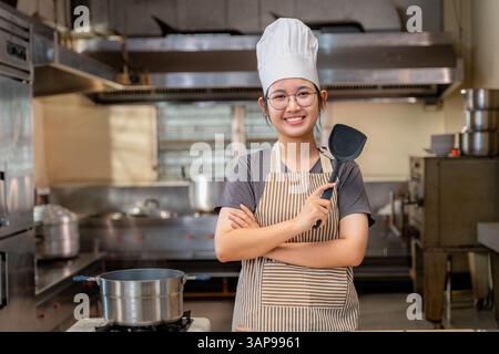 Smiling young female chef in professional kitchen attire, holding a spatula and standing proudly behind a counter filled with fresh vegetables and coo Stock Photo