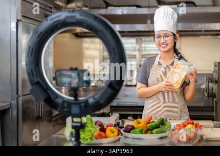 A young female chef wearing a striped apron and white chef hat records a cooking tutorial using a smartphone and ring light setup in a commercial kitc Stock Photo