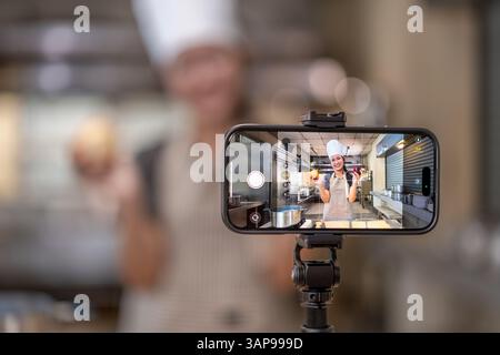 A smartphone mounted on a tripod records a young female chef in a professional kitchen. The camera screen shows the chef smiling and holding vegetable Stock Photo