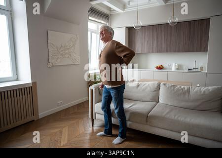 Sad elderly man painfully straightens hunched back after rising from couch with severe spinal pain Stock Photo