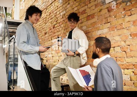 Three team members collaborate in a vibrant office, sharing ideas over coffee and tech. Stock Photo