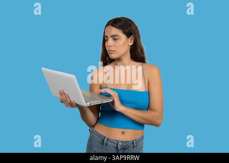 Focused young woman using a laptop against a vibrant blue backdrop Stock Photo