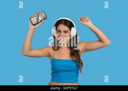 Happy young woman wearing headphones listening to music on blue background, holding smartphone and dancing Stock Photo