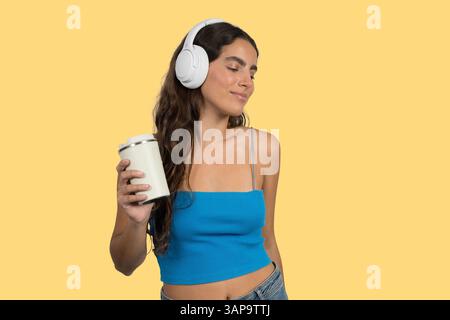 Carefree young woman holding reusable coffee cup while listening music with wireless headphones, isolated on yellow background Stock Photo