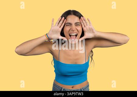 Young woman wearing blue top and rainbow bracelet is framing her face with hands and screaming on yellow background Stock Photo