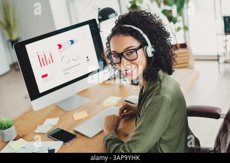 Young professional woman wearing headphones working in a modern office setting, analyzing data charts on a large computer monitor Stock Photo