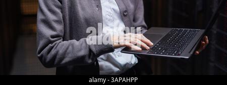 Website header shot of person wearing cardigan with tattooed hands typing on laptop in data center setting surrounded by servers and equipment Stock Photo