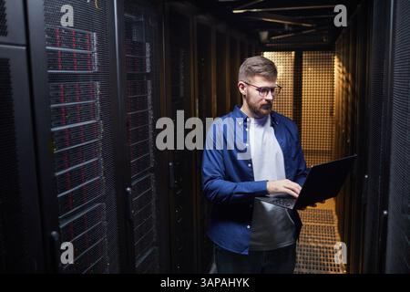 IT professional working in a server room managing hardware and software with focused expression. Surrounded by server racks filled with equipment, creating tech-focused environment Stock Photo