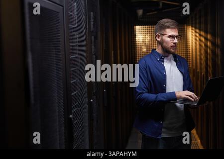 Man operating laptop in data center surrounded by server racks and equipment. Concentrated professional monitoring network systems and ensuring smooth operations Stock Photo