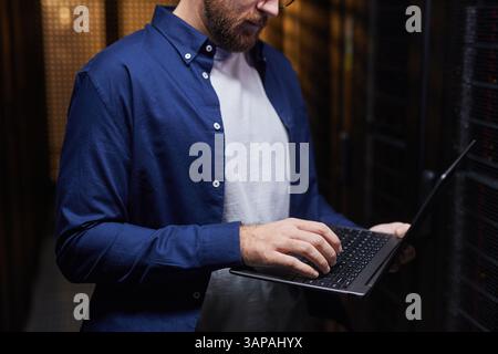 Man focusing on his laptop in a data center, surrounded by server racks, concentrating on his task in technology-driven environment Stock Photo