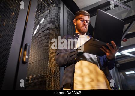 Man inspecting server rack while holding laptop in modern data center with focused expression. Image shows male professional in technology-rich setting Stock Photo