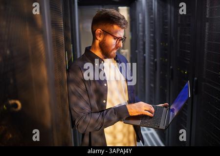 Portrait of a Caucasian male technician working in a data center checking a server on a laptop. Reflecting focused and concentrated expression while standing in server room Stock Photo