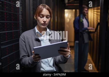 Woman working on tablet in data center with man in background using laptop. Focused on her tasks, highlighting modern technological environment and concentration on responsibilities Stock Photo