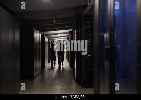Technicians silhouetted against the light while walking through a server room corridor, surrounded by towering racks of servers, discussing and inspecting equipment Stock Photo